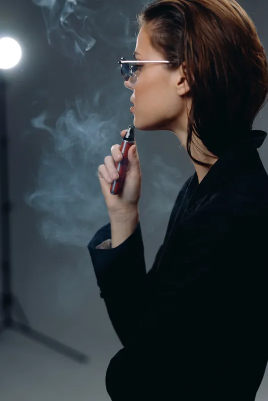 Stylish woman vaping in dark indoor setting with smoke and modern lighting, wearing glasses and black jacket.