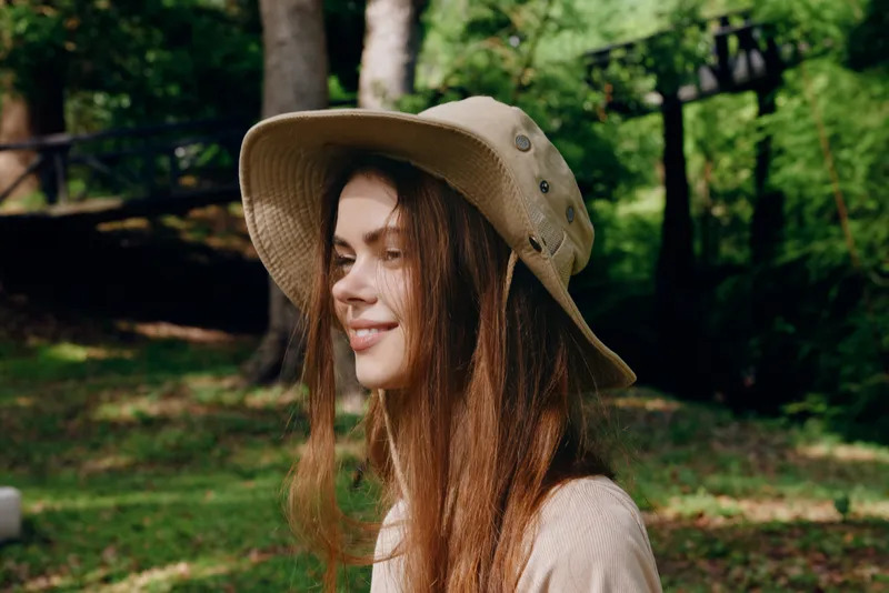 Woman portrait smile hat outdoors nature in a sunlit park, side profile closeup with long hair and relaxed expression, casual outfit and soft natural lighting for lifestyle imagery.