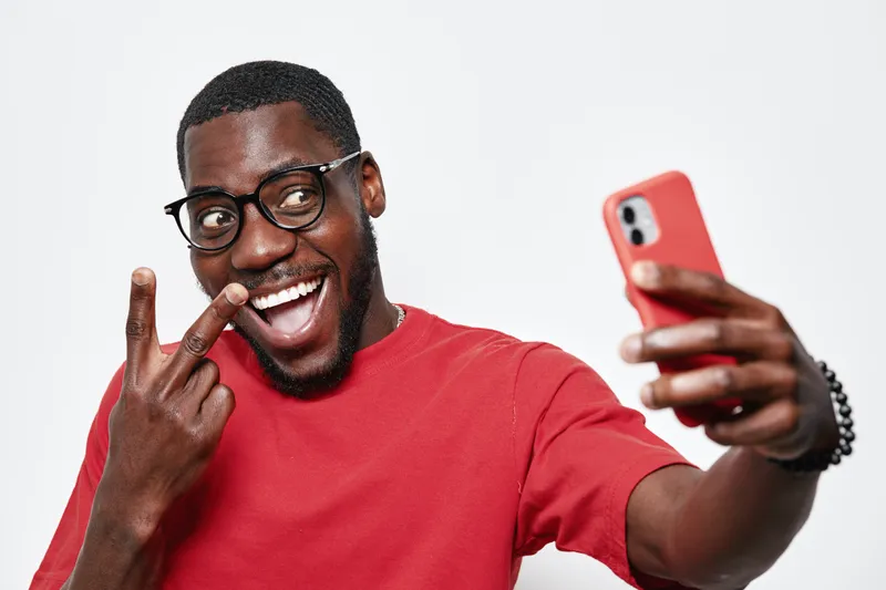 African American man with dark skin and glasses taking selfie with smartphone smiling and showing peace sign in red shirt on white background. Happy young male portrait.