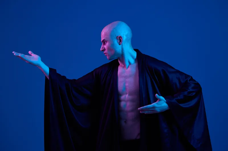 Male performer in a flowing black robe, showcasing a dramatic pose under blue lighting. The image emphasizes movement and expresses creativity in a unique style.