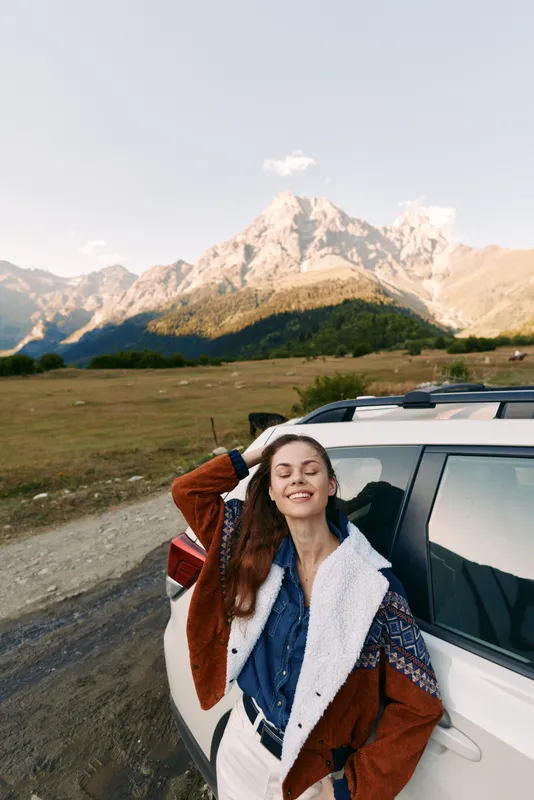 Woman car mountains nature travel road smile jacket outdoors adventure. Young woman leans on white SUV on a scenic mountain road, smiling with eyes closed in cozy shearling jacket, relaxed travel