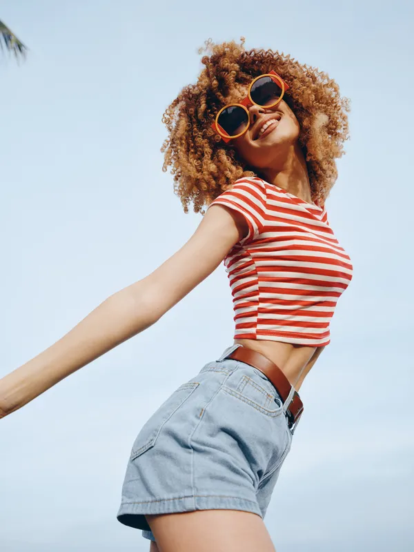 Joyful young woman with curly hair wearing a red striped crop top and denim shorts, posing under a clear blue sky, exuding carefree summer energy and style.