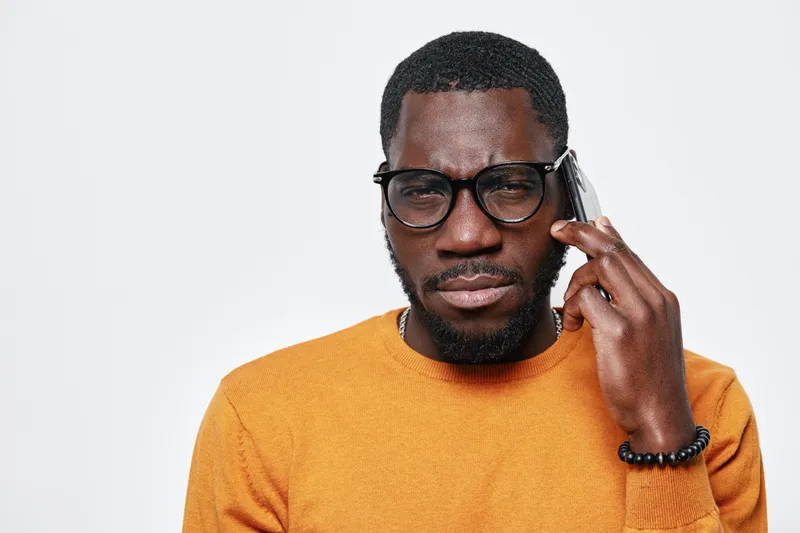 African American man with dark skin wearing glasses and orange sweater talking on smartphone with serious expression isolated on white background. Communication and technology concept.