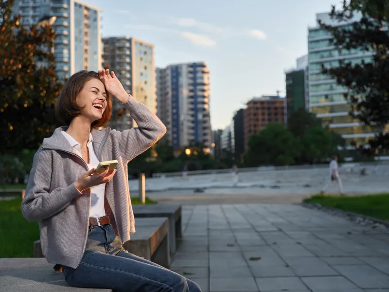 A cheerful young woman sits on a park bench in an urban setting, texting on her smartphone. She smiles brightly, enjoying a relaxed moment amid modern high rise buildings and clear daylight.