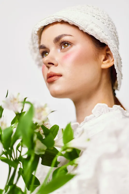 Young woman in a white hat holding flowers, showcasing natural beauty and soft pastel colors, exuding serenity and grace in a bright studio setting