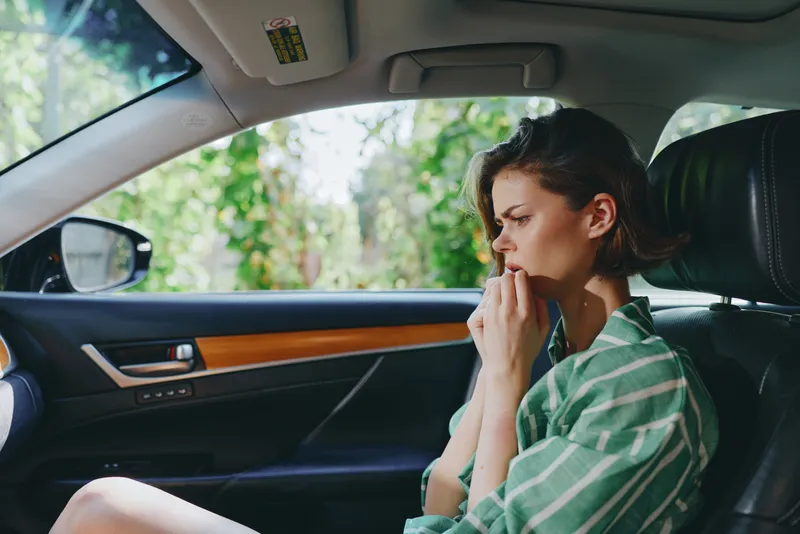 A thoughtful woman sits in a car interior, touching her lips with a worried, anxious expression as she gazes forward.