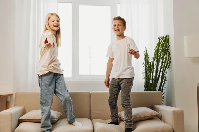 Happy children jumping on beige sofa in bright living room with large window and green plant. Concept of playful kids, home fun, and family time.