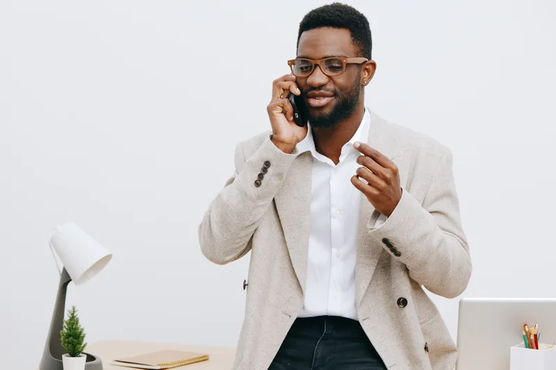 Professional shot of a smiling man with dark skin and glasses talking on a mobile phone while standing in an office environment, wearing a light blazer and white shirt, with a desk, laptop, and plant 