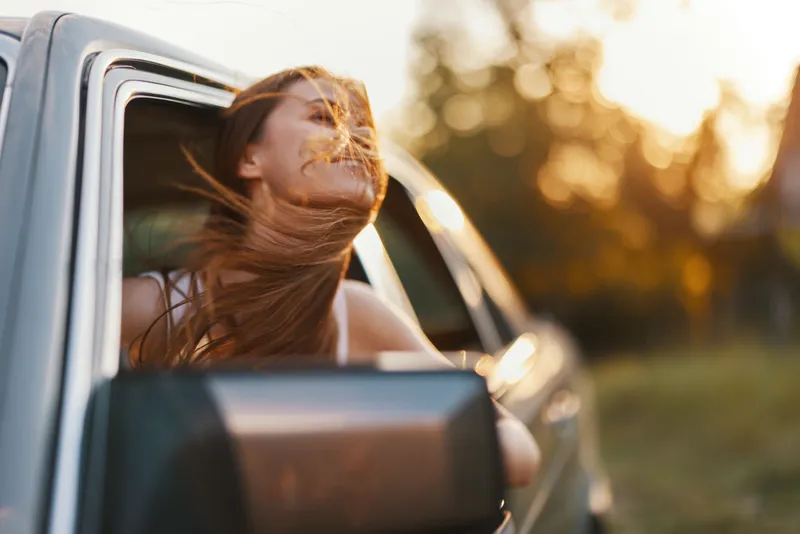 Young woman enjoying a carefree moment with flowing hair out of the car window during sunset, capturing the essence of freedom and joy.