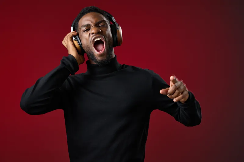 Young Black man wearing headphones, singing joyfully against a red background, showcasing passion and enjoyment of music KEY music, headphones, singing, joy, emotion, red, young, male, passion, hap