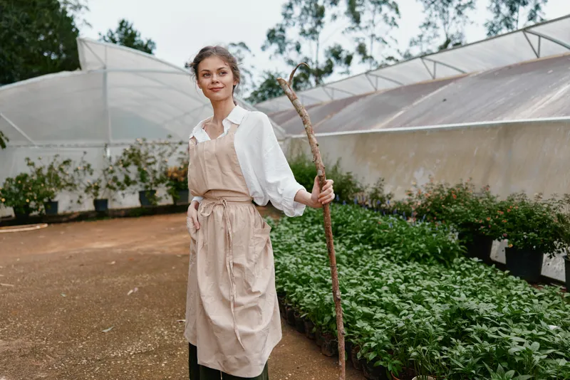 Young woman in an apron smiling joyfully in a lush greenhouse, holding a stick, surrounded by vibrant plants and greenery, showcasing a connection to nature