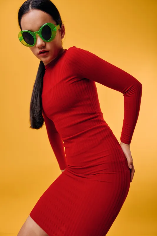 Young woman in red dress posing on yellow background with long hair and green sunglasses. Fashion portrait with bold colors and dynamic pose showcasing modern style and attitude.