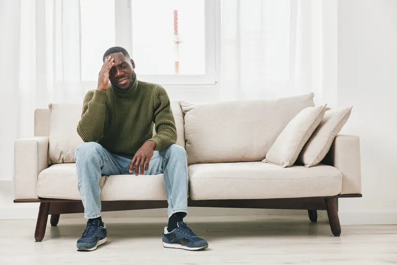 Emotional African American man sitting on a sofa, expressing sadness while wearing a sweater, jeans, and sneakers in a bright, minimalistic room
