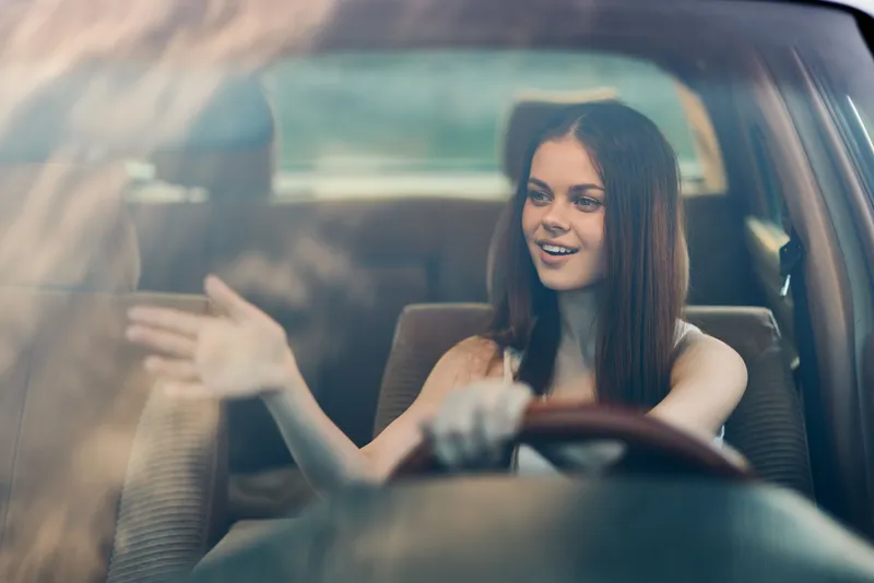 Happy young woman driving a car, waving from the window with a joyful smile, showcasing vibrant emotions and a sunny atmosphere.