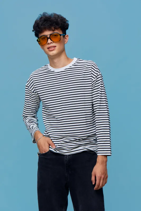 Young man wearing striped long sleeve shirt and sunglasses poses confidently against blue background with casual style and trendy look in modern fashion portrait.