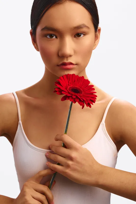 Asian woman holds a bold red flower near her chest in a simple studio portrait. She wears a white tank top, with calm expression and soft lighting against a clean background, creating an elegant,