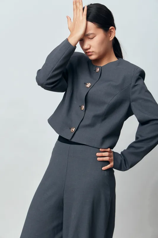 Woman in gray suit with closed eyes holds hand on forehead standing against light background, showing stress and fatigue. Business style, female model, indoor portrait with emotion.