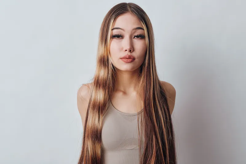 Young woman with long straight hair posing in beige tank top against neutral background in studio. Female model looking at camera with confident expression and natural makeup.