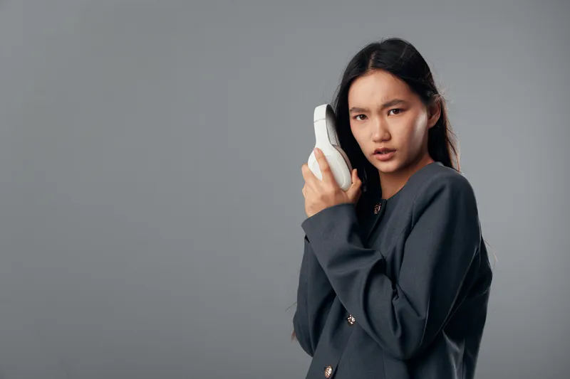 Young woman holding white headphones near ear with serious expression against gray background. Female model in dark jacket posing with audio accessory for music or communication concept.