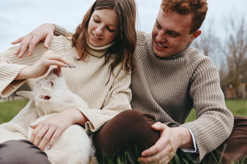 Smiling couple in cozy sweaters gently strokes a white goat kid in a grassy yard, capturing a warm lifestyle moment. Natural light, candid interaction, animals, harmony, tenderness, companionship,