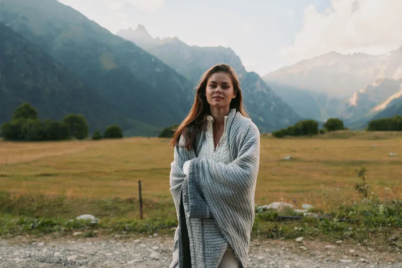 Woman in a cozy shawl stands in a grassy field with distant mountains, showcasing outdoor lifestyle, calm self care, and nature inspired serenity, and warm tones create inviting mood.