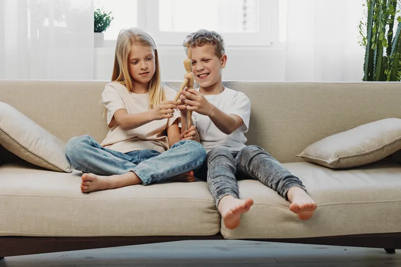 Two children sitting on a beige sofa playing with wooden toys in a bright living room with natural light and casual clothing.