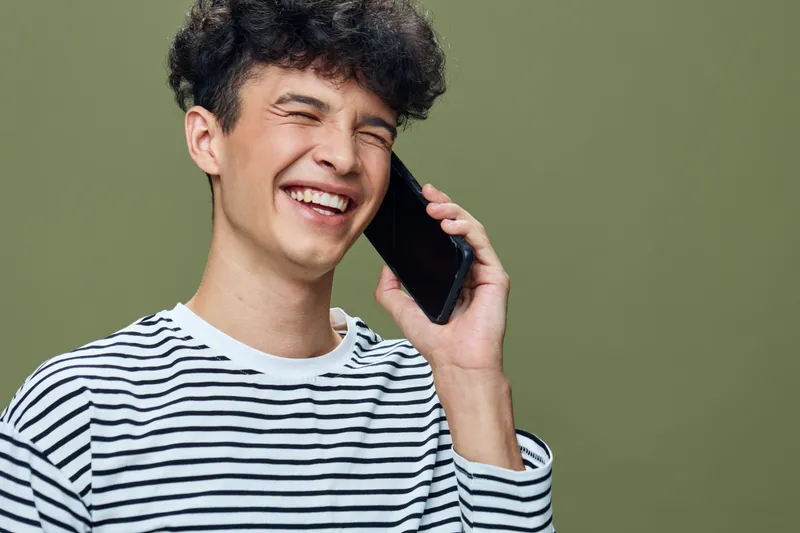 smiling man talking on phone wearing striped shirt with curly hair, expressing happiness and amusement against plain olive background.