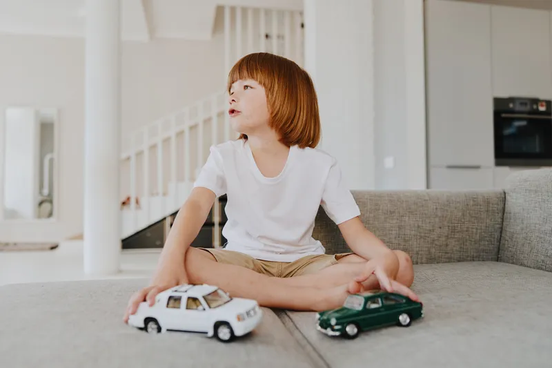 A young boy sitting on a sofa, playing with toy cars in a bright, modern living room. His joyful expression highlights the fun of childhood and imaginative play.