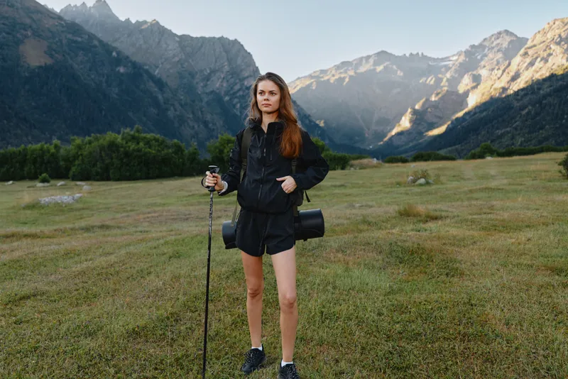 A solo hiker stands in a rugged mountain landscape, equipped with trekking poles and a camera bag. She looks confident and ready for adventure amid vast scenery and dramatic peaks.