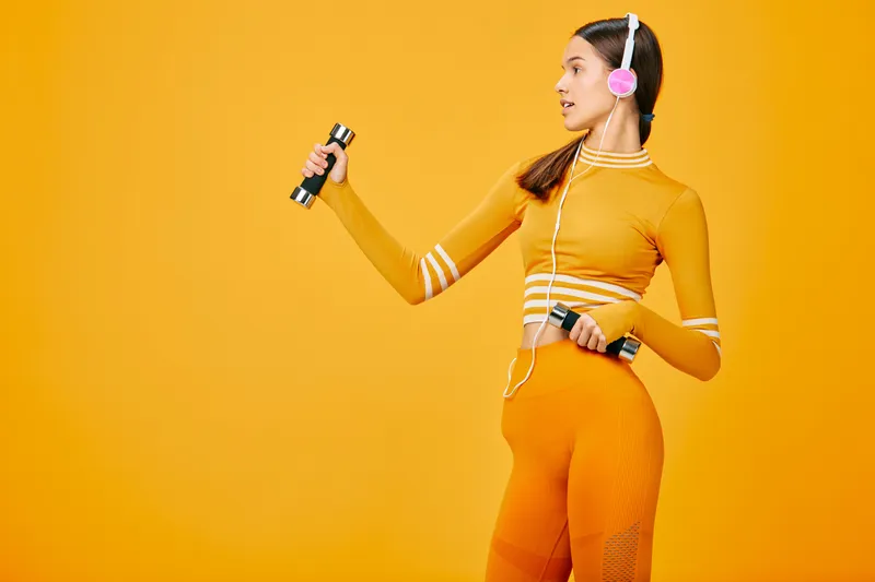 Fitness woman in a bright orange athletic set lifts dumbbells in a dynamic workout scene, wearing headphones, showcasing energy, strength, and sport focus in a vibrant studio.