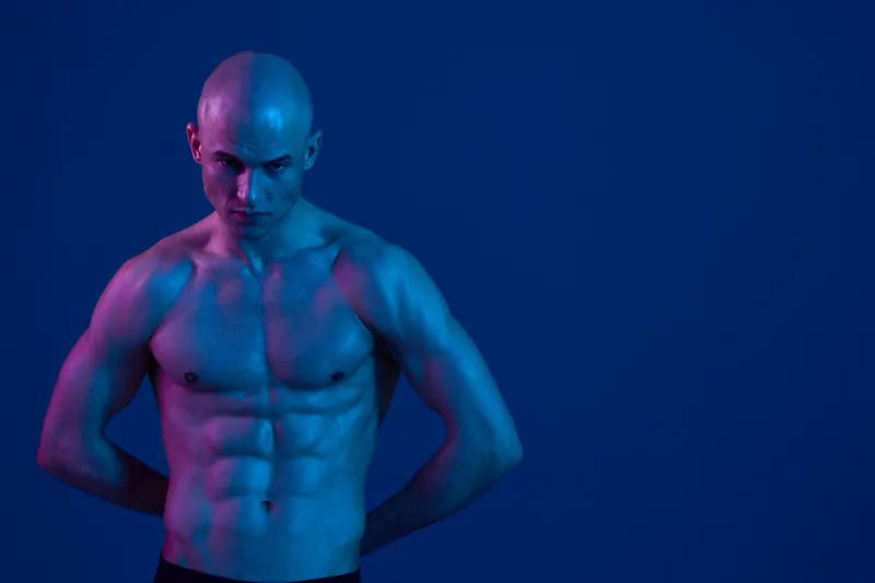 Muscular man posing in low light with a blue background, showcasing strength and determination in a captivating black and white style.