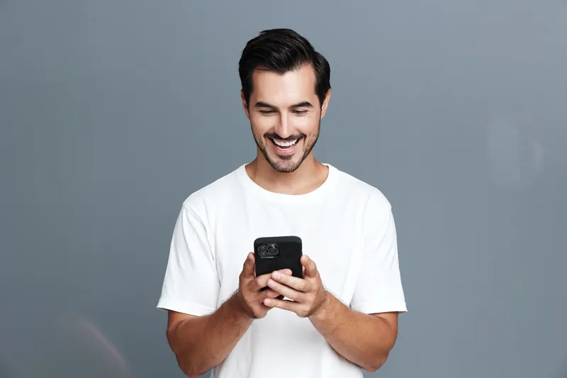 Smiling young man wearing a white t-shirt, looking at his mobile phone with a joyful expression against a plain gray background. People technology lifestyle concept.