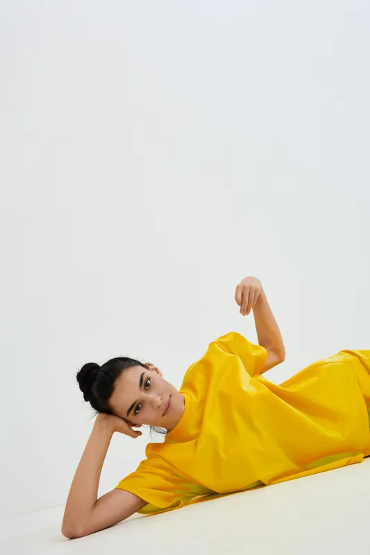 Young woman in bright yellow t-shirt lying on the floor, relaxed pose and thoughtful expression in minimalistic white space, casual clothing indoors.
