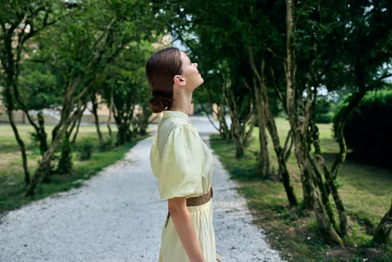 A woman stands in a sunlit park along a gravel path, wearing a pale dress and belt, gazing upward with calm and a sense of discovery in a lush natural setting.