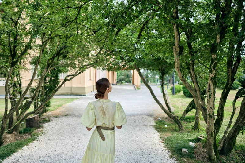 Woman walking along a gravel path in a lush garden, wearing a pale dress, seen from behind, creating a serene outdoor scene with soft light and greenery.
