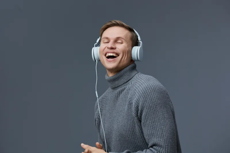 Smiling young caucasian man wearing gray turtleneck sweater listening to music with large white headphones on plain gray background studio portrait, expression of joy, happiness, leisure, relaxation, 