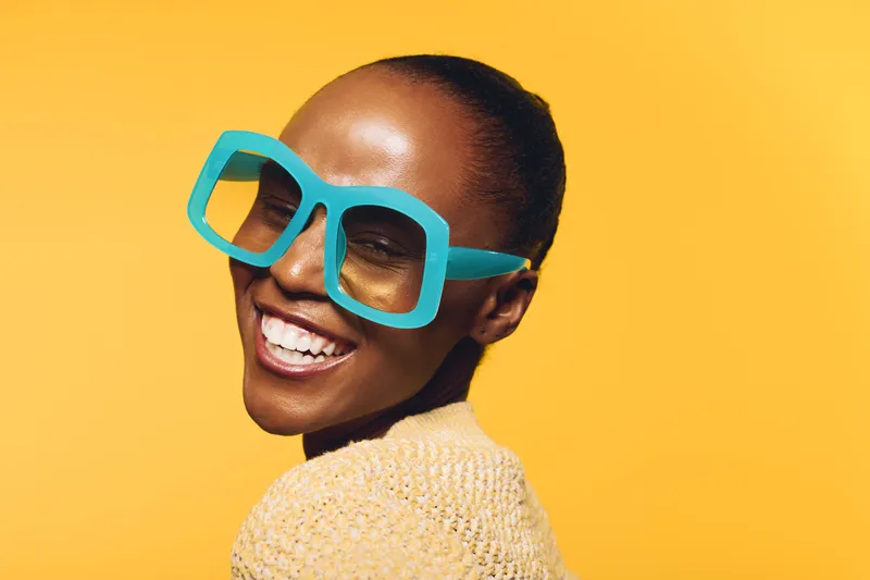 Smiling African American woman with dark skin wearing oversized blue sunglasses and a yellow sweater poses against a bright yellow background. Concept of fashion, joy, and summer style.