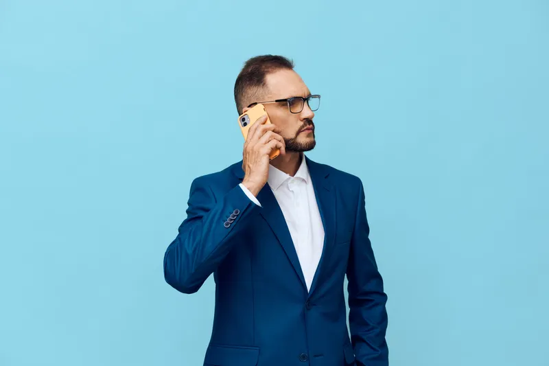 An urban professional man in a blue suit speaks on a smartphone against a solid blue background, projecting focus, confidence, and calm in a studio portrait.