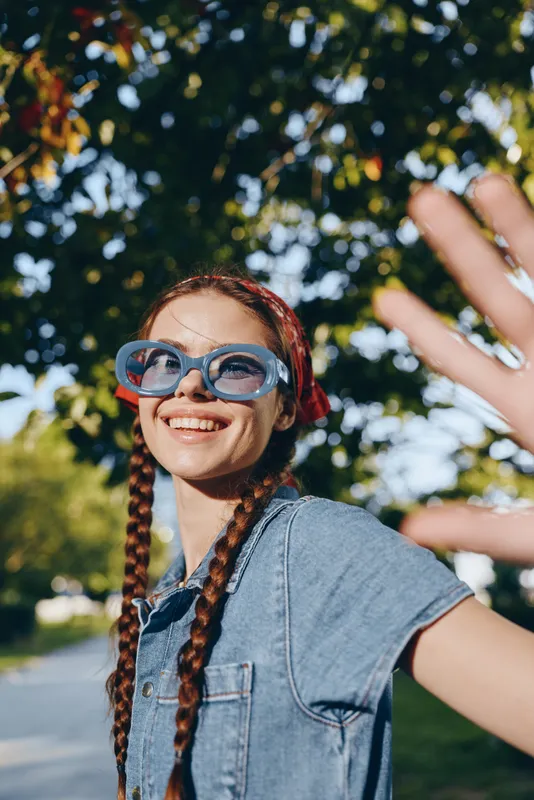 smiling woman with braids wearing blue sunglasses and denim shirt outdoors with green trees and sunny weather smiling joyfully