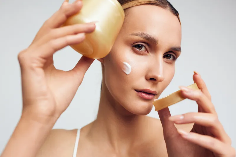Woman applying cream on face holding open jar with natural skincare product. Close-up portrait with soft lighting on neutral background. Concept of beauty, self-care, and wellness.