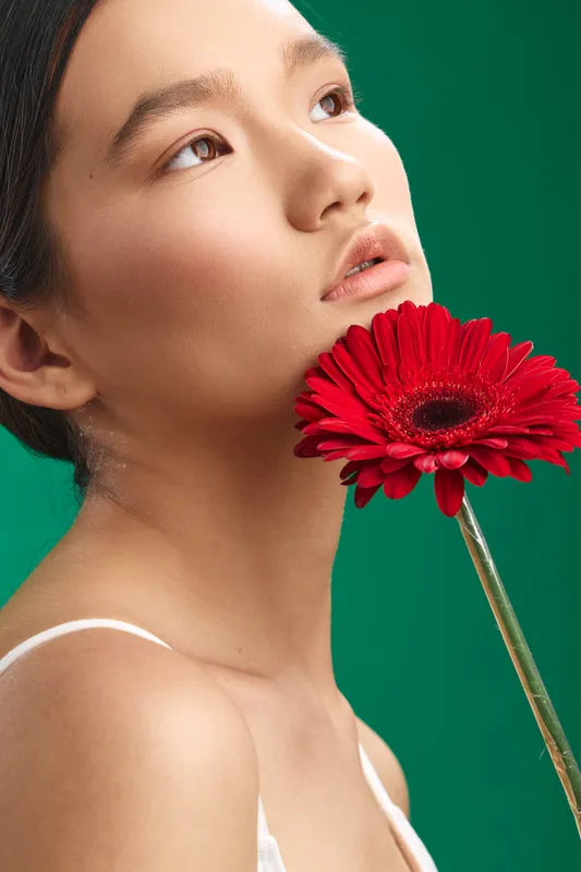 asian woman gazes softly at a red flower held close in front of a vibrant green background, conveying calm beauty and an intimate portrait mood.