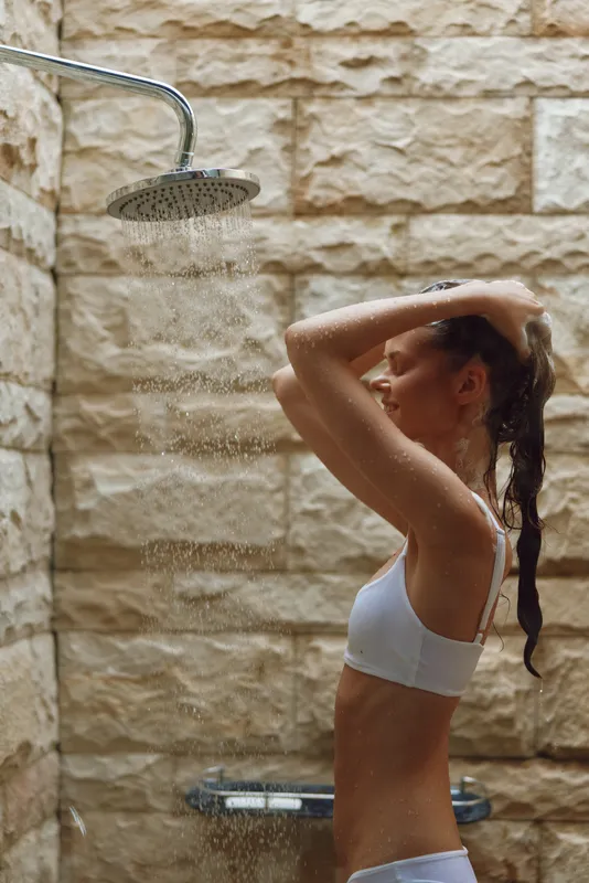 woman taking shower under rainfall showerhead in modern stone wall bathroom, natural light highlighting wet skin and relaxed expression