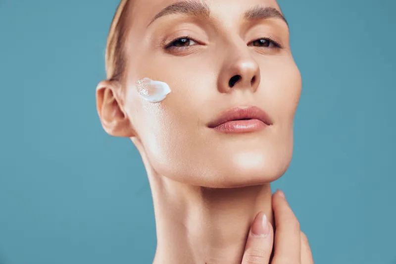 Woman face close up, skincare beauty routine, white cream on cheek, hand on chin, smooth glowing skin, fresh spa feeling, soft studio light, blue background
