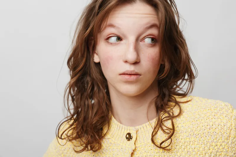 Young woman with curly brown hair wearing yellow sweater looks sideways with curious expression on light grey background. Concept of emotion, curiosity, and casual portrait.