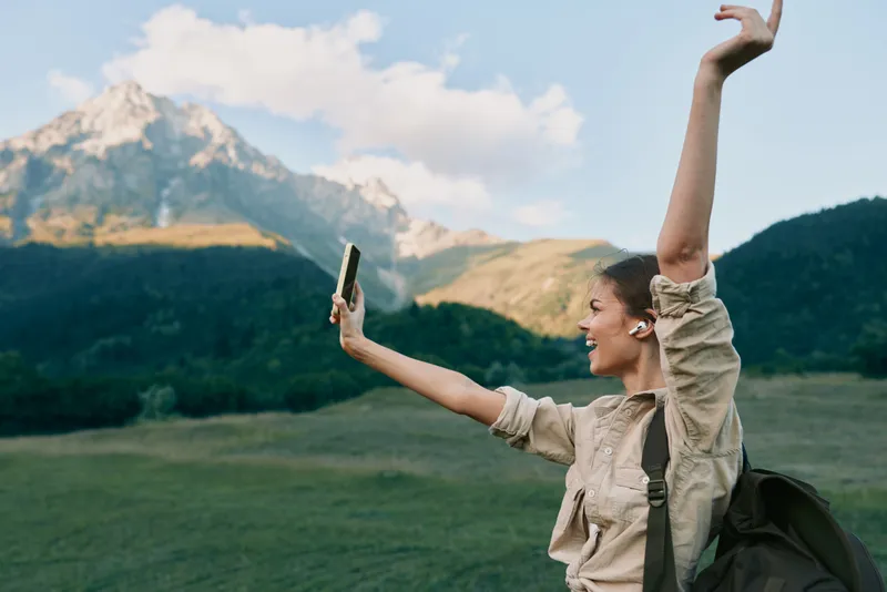 Young woman with a backpack takes a selfie with a smartphone while smiling and raising her arm in front of a mountain landscape during daytime.