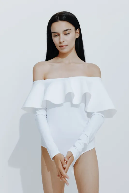 young woman sport fitness wearing white off-shoulder bodysuit with long gloves standing against bright wall, minimalistic style, serene expression and natural light