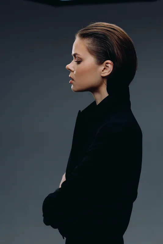 Profile of a thoughtful woman in black clothing with a minimalist dark background and moody lighting