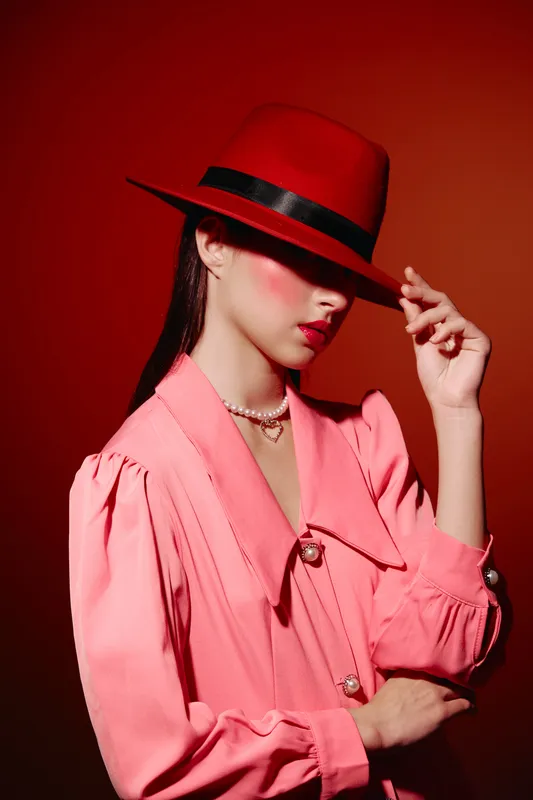 Stylish woman in a red hat and pink jacket covers her face while posing against a vivid red background. Creative beauty and fashion portrait with dramatic lighting.