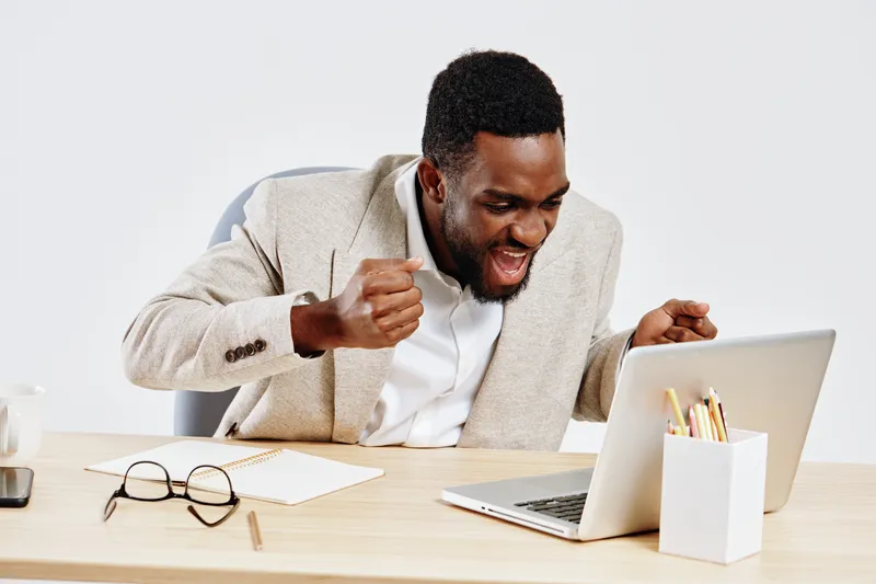 Smiling man with dark skin celebrating success while looking at laptop screen in modern office setting, expressing happiness and excitement, business achievement, workspace, technology, positive emoti
