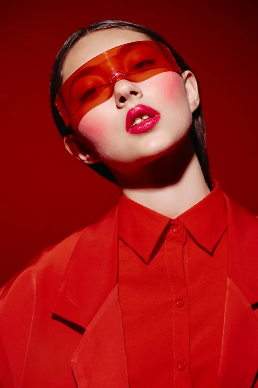 Fashion model with wet slicked back hair wears red transparent sunglasses and bright lipstick. Red coat and shirt on red gradient backdrop. Creative beauty editorial concept.